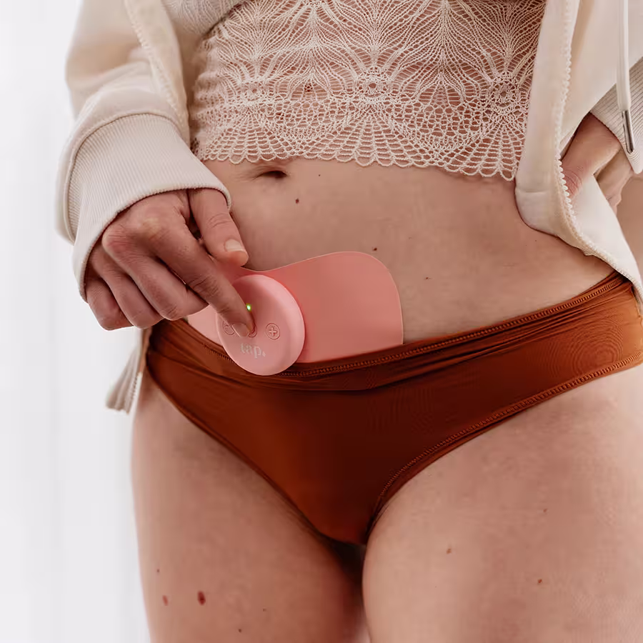 A woman wearing a pink Tap wearable device on her lower abdomen, pressing a button on the circular controller. She wears a lace top, cream hoodie, and rust-coloured underwear.